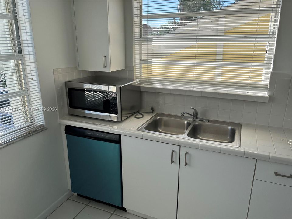Kitchen featuring white cabinetry, tile countertops, dual-basin stainless steel sink, tiled backsplash, and built-in dishwasher