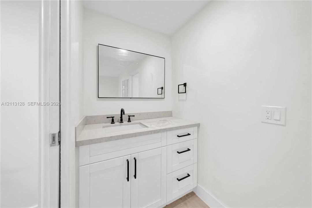 Modern vanity featuring white shaker-style cabinetry, matte black hardware, a light-toned stone-finish countertop, integrated basin, and a framed rectangular mirror
