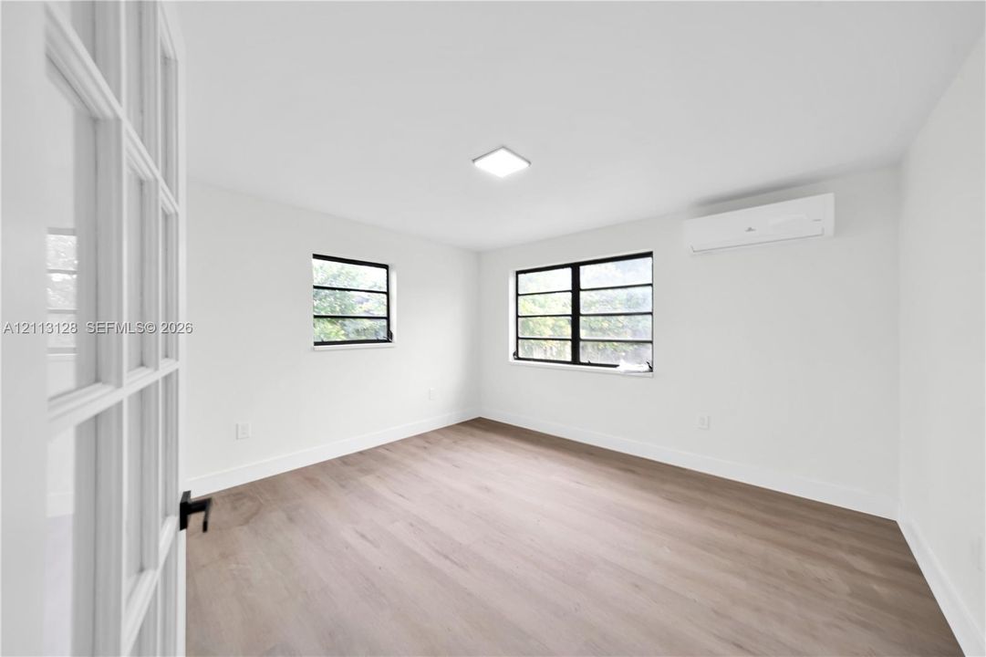 Interior space featuring wood-finish flooring, windows with black frames, wall-mounted ductless climate control unit, and a glass-paneled entry door