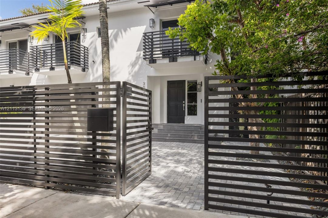 Exterior facade featuring a paved stone driveway, horizontal slat metal privacy fencing, solid panel entry door, and upper-level balconies with matching metal railings