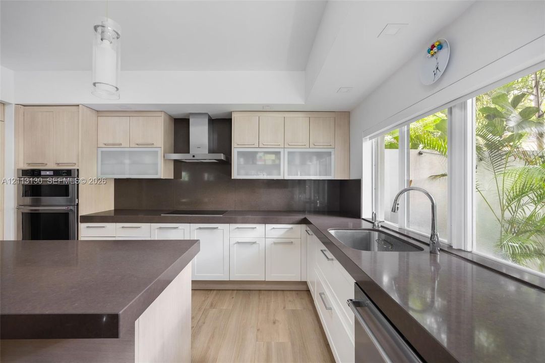Kitchen featuring light wood-finish cabinetry, solid-surface countertops, a stainless steel chimney-style range hood, built-in double ovens, and a dual-aspect window