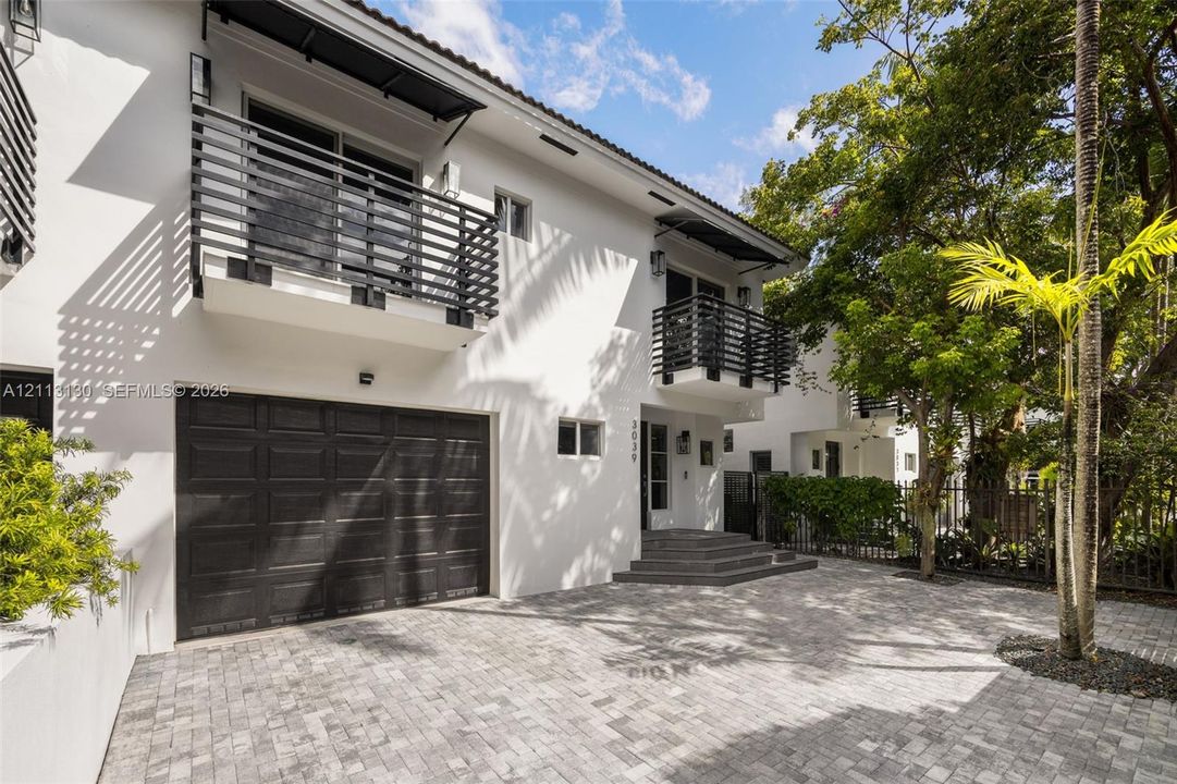 Exterior featuring smooth white stucco siding, dual second-story balconies with horizontal slat railings, a paved stone driveway, and an attached multi-car garage with dark paneled doors
