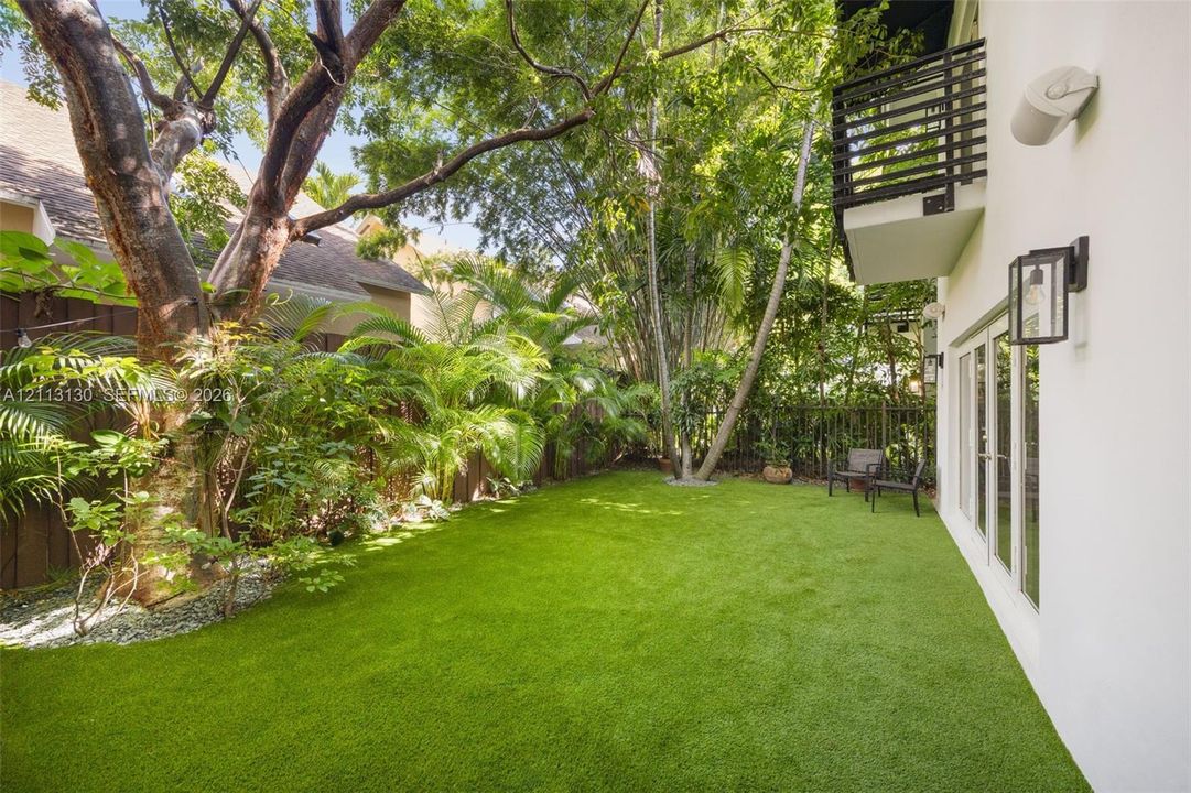 Private backyard featuring synthetic turf, mature trees, wood privacy fencing, a second-story balcony with metal railings, and exterior wall-mounted lanterns