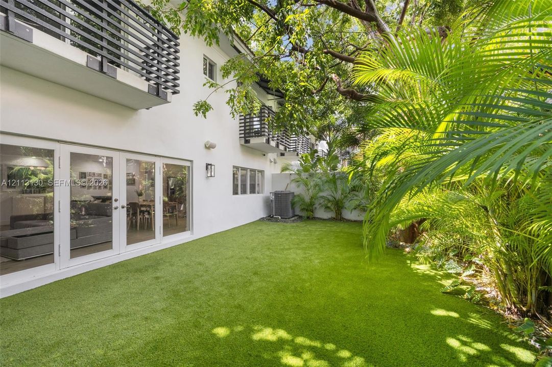Backyard featuring artificial turf, white stucco exterior, multiple second-story balconies with horizontal railings, and sliding glass panel doors