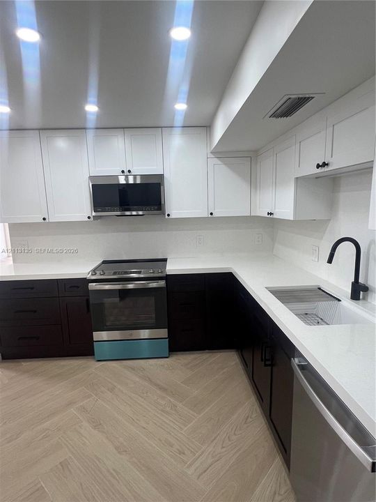Kitchen featuring two-tone cabinetry, white stone-composite countertops, stainless steel appliances, matte black hardware, and wood-finish herringbone flooring