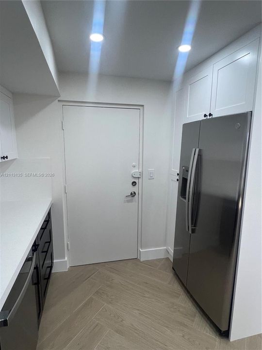 Shaker-style white cabinetry, stainless steel side-by-side refrigerator, solid-surface countertops, recessed lighting, and herringbone-patterned flooring