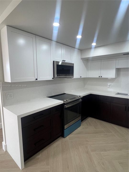 Two-tone cabinetry featuring white upper cabinets and dark lower drawers, solid surface countertops, stainless steel over-the-range microwave, electric range, and wood-finish flooring in a herringbone pattern
