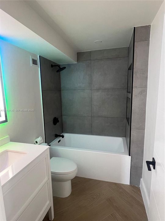Bathroom featuring large-format gray tile shower surround with integrated shelving, white vanity with rectangular basin, matte black fixtures, and herringbone-patterned wood-finish flooring