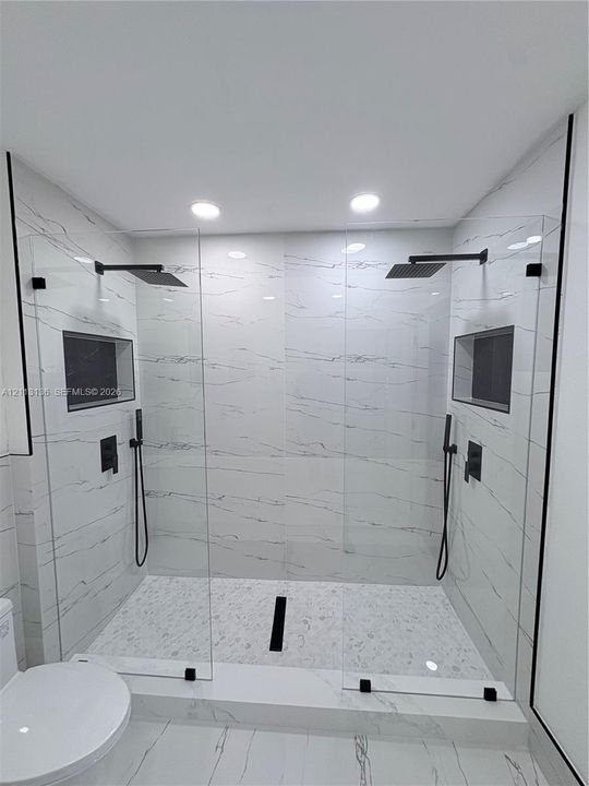 Walk-in shower featuring large-format stone-patterned tile walls, dual rainfall showerheads, handheld sprayers, built-in recessed niches, and matte black hardware
