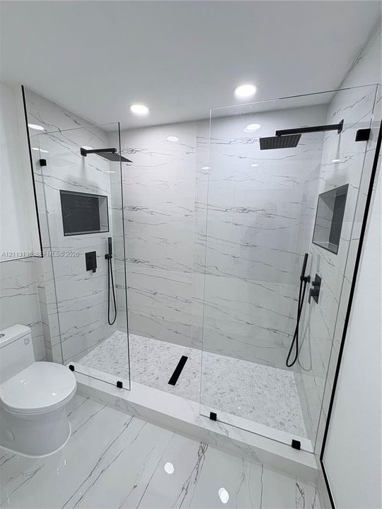 Walk-in shower featuring dual shower heads with matte black fixtures, frameless glass enclosures, built-in recessed shelving, and stone-finish wall tile