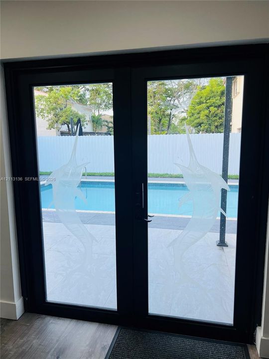 Black-framed double glass doors featuring custom frosted marlin etchings, opening to a pool deck with paved surfaces and perimeter fencing