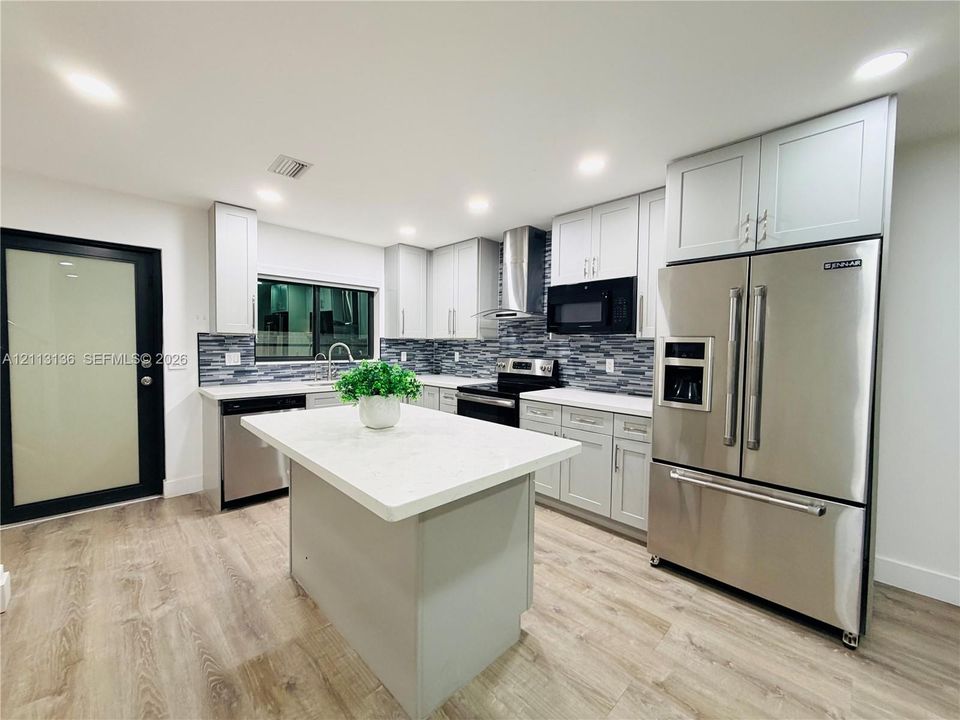 Kitchen featuring light gray shaker-style cabinetry, quartz countertops, a central island, stainless steel appliances, and a glass-tiled backsplash