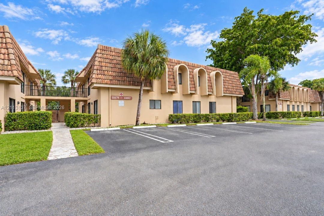 Two-story exterior featuring a clay tile roof, arched architectural accents, beige stucco finish, designated surface parking, and manicured perimeter landscaping