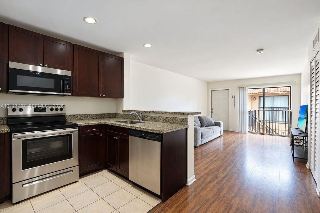Kitchen featuring dark wood cabinetry, stone-finish countertops, stainless steel appliances, and tile flooring, paired with wood-finish flooring and a private balcony