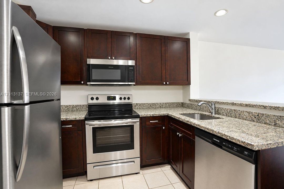 Kitchen featuring dark wood cabinetry, stone-patterned countertops, stainless steel appliances, a recessed sink, and tile flooring