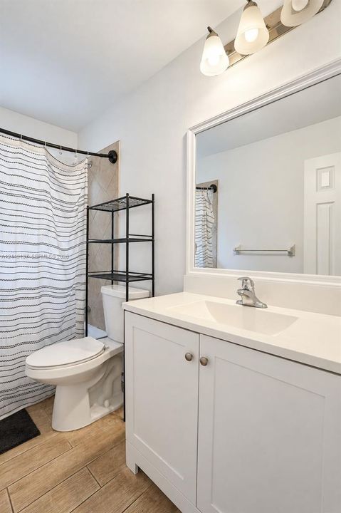 White vanity with a solid-surface countertop and integrated sink, framed wall mirror, three-light vanity fixture, and wood-finish flooring