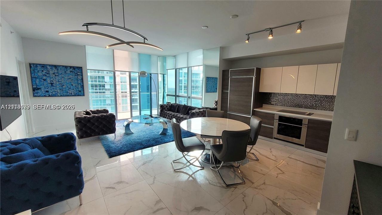 Open-concept living space featuring floor-to-ceiling windows, large-format tile flooring, modern kitchen cabinetry with built-in appliances, and designer-style light fixtures