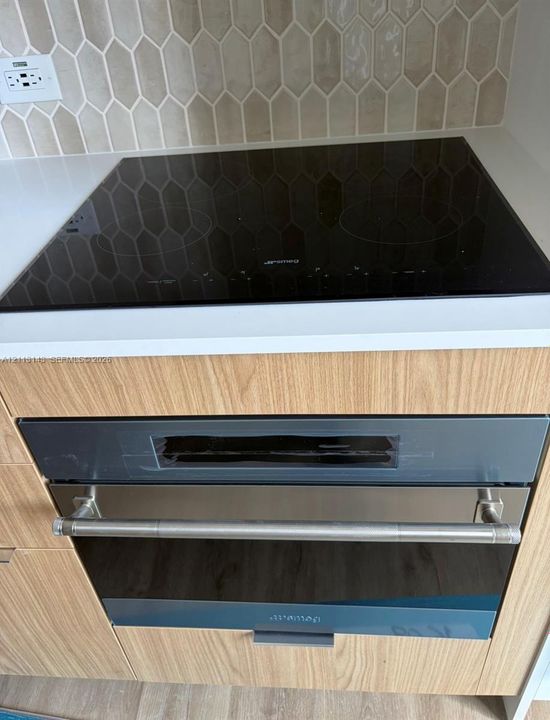 Built-in Smeg induction cooktop and matching wall oven with stainless steel handle, set within light wood-finish cabinetry, solid surface countertops, and a decorative hexagonal tile backsplash