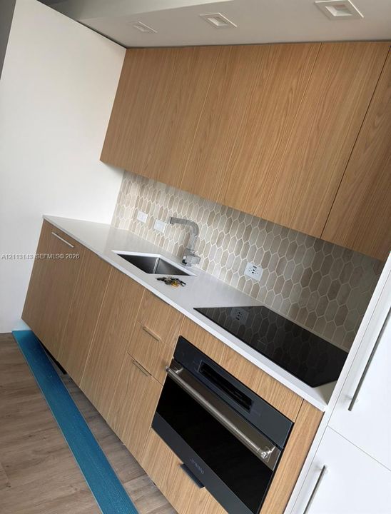 Kitchen featuring wood-finish cabinetry, white solid-surface countertops, a textured hexagonal tile backsplash, a stainless steel sink with a single-handle faucet, and a built-in cooktop and oven