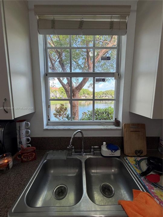 Double-basin stainless steel sink with a high-arch faucet, flanked by white cabinetry and stone-finish countertops, featuring a multi-pane window with a water-facing orientation