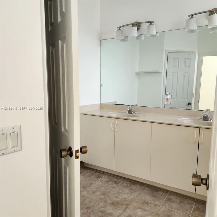 Bathroom featuring a dual-sink vanity with solid-surface countertops, expansive wall-to-wall mirror, multi-bulb light fixtures, and neutral floor tile