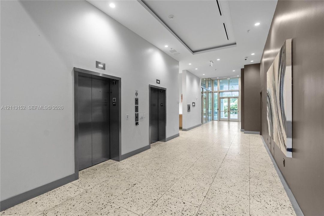 Elevated lobby featuring dual stainless steel elevators, large-format speckled stone flooring, recessed ceiling lighting, and floor-to-ceiling glass entry doors
