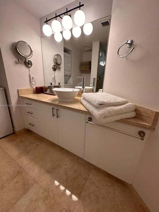 Vanity featuring a vessel sink, stone countertop, white cabinetry with metal hardware, large wall-mounted mirror, and multi-bulb overhead lighting