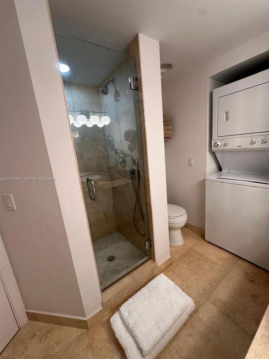 Full-height frameless glass shower enclosure with tiled interior, integrated stackable laundry unit, and stone-finish floor tiling