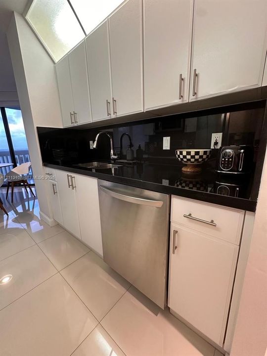 Kitchen featuring white cabinetry with metallic pulls, dark solid-surface countertops, a stainless steel dishwasher, a sleek gooseneck faucet, and light-toned tile flooring