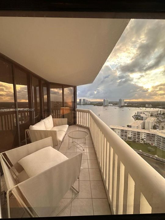 Covered balcony featuring expansive glass paneling, tile flooring, and a white vertical railing, overlooking an expansive water landscape and urban skyline