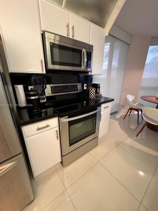 Kitchen featuring white shaker-style cabinetry, polished dark stone countertops, stainless steel appliances, and large-format ceramic tile flooring
