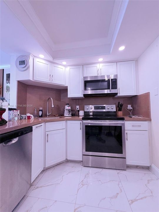 Kitchen featuring white shaker-style cabinetry, stainless steel appliances, solid surface countertops, a high-arch gooseneck faucet, and large-format flooring with a veined pattern