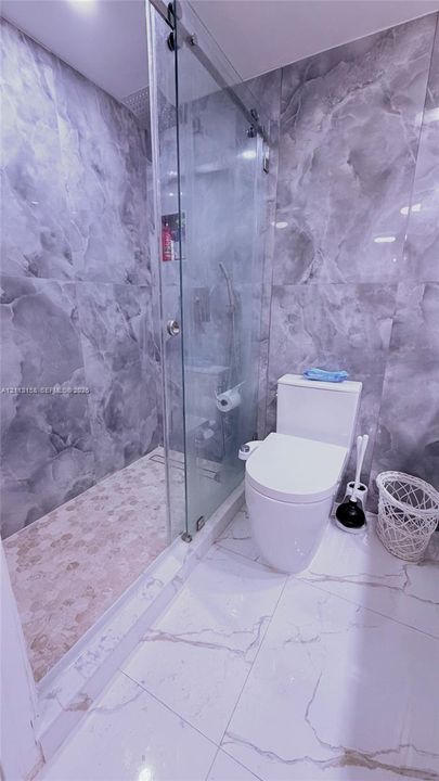 Walk-in shower with a sliding glass door, floor-to-ceiling gray-toned wall tiling, pebble-finish shower flooring, and oversized white floor tile with gray veining