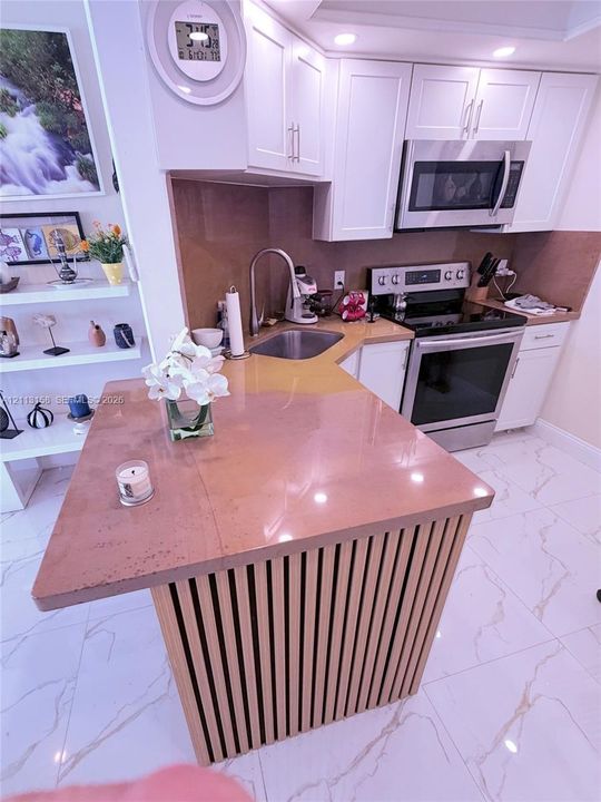 Kitchen featuring a slatted wood-finish island base, solid-surface countertop, white shaker-style cabinetry, stainless steel appliances, and tile flooring