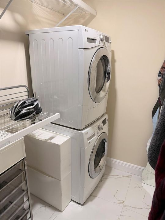 Stacked front-load laundry appliances, white stone-finish tile flooring, and integrated white wire shelving