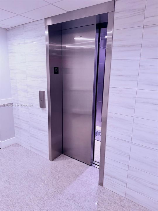 Stainless steel elevator with a brushed metal frame, surrounded by large-format, horizontal-vein tile wall cladding and a polished stone-finish floor