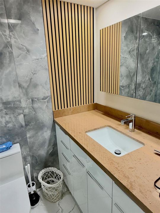 Custom vanity featuring a solid surface countertop with an integrated basin, high-gloss white cabinetry, vertical wood-slat accent wall, and large-format gray patterned wall tile