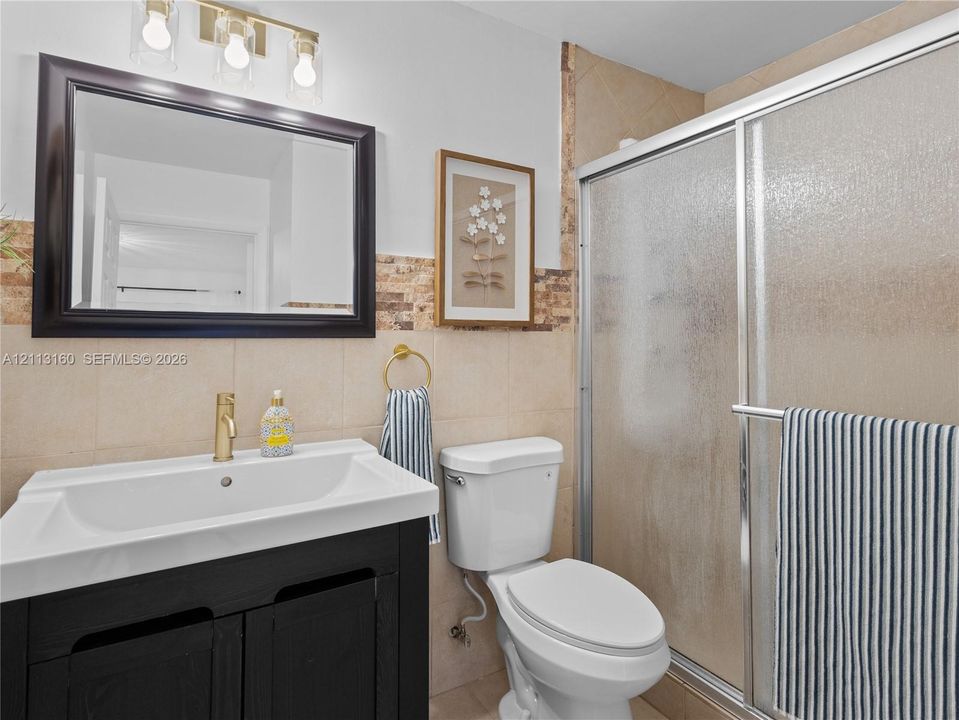 Dark-finish vanity with integrated rectangular sink and gold-tone faucet, framed wall mirror, three-light wall-mounted fixture, walk-in shower with sliding glass doors, and natural stone tile backsplash