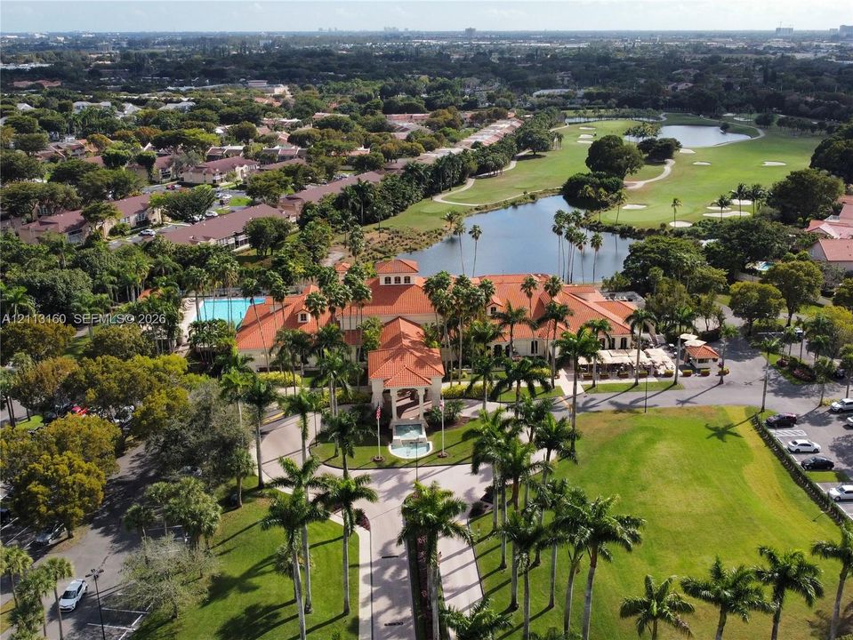 Aerial view featuring a Mediterranean-style clubhouse with a terra-cotta tile roof, landscaped grounds with mature palm trees, a swimming pool, and proximity to a golf course with water features