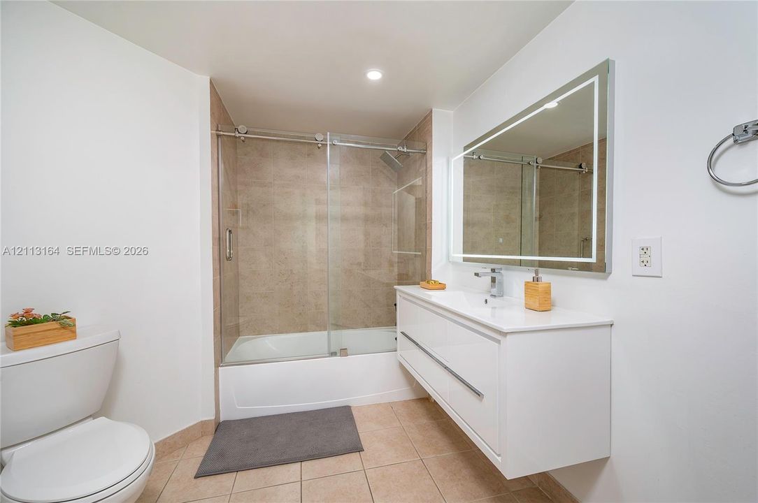 Floating white vanity with a rectangular sink, wall-mounted rectangular mirror with integrated LED lighting, and a glass-enclosed tub shower with tile surround