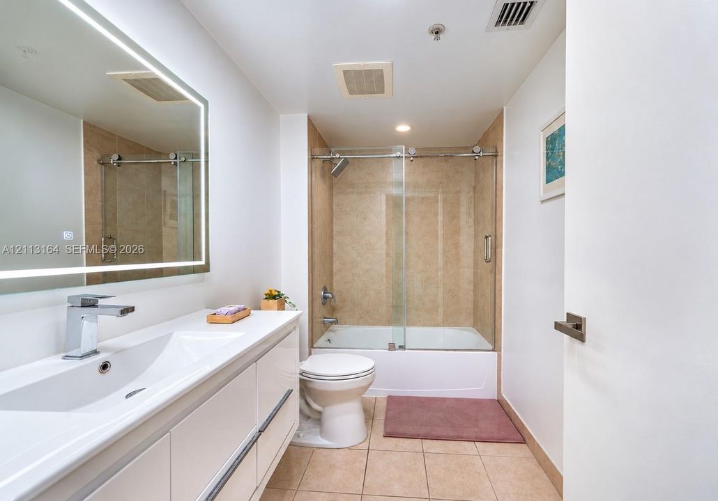 Modern bathroom featuring a floating white vanity with integrated sink, backlit vanity mirror, ceramic tile flooring, and a tub-shower combination with a glass sliding door
