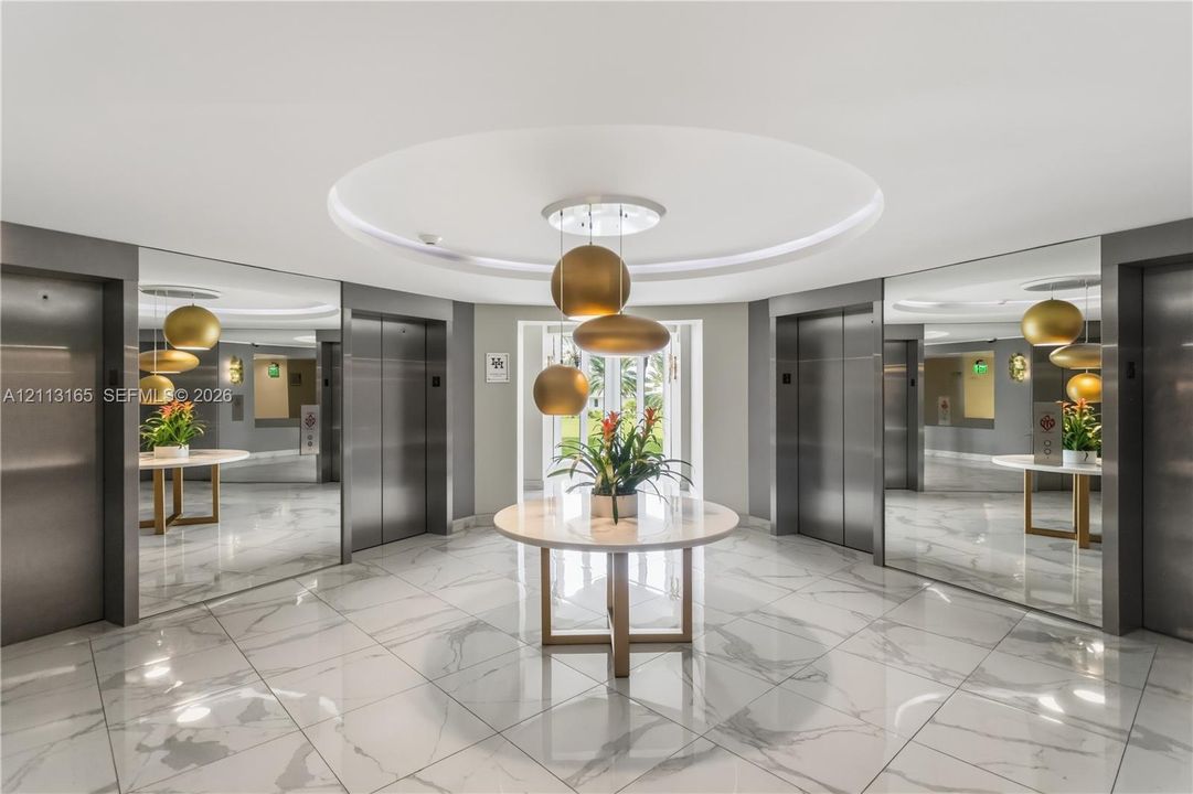 Elevator lobby featuring polished stone flooring, recessed circular ceiling detail, stainless steel elevator doors, full-height wall mirrors, and tiered metallic pendant lighting