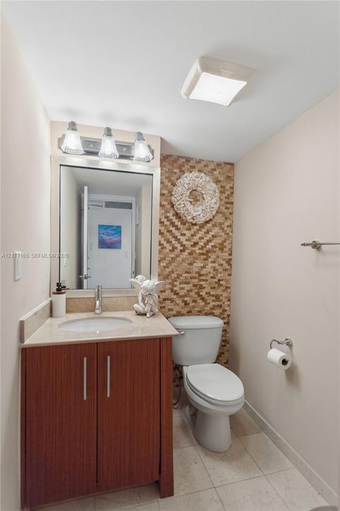 Vanity with a solid-surface countertop and integrated sink, wood-finish cabinetry, mosaic-tiled accent wall, three-light fixture, and ceramic tile flooring