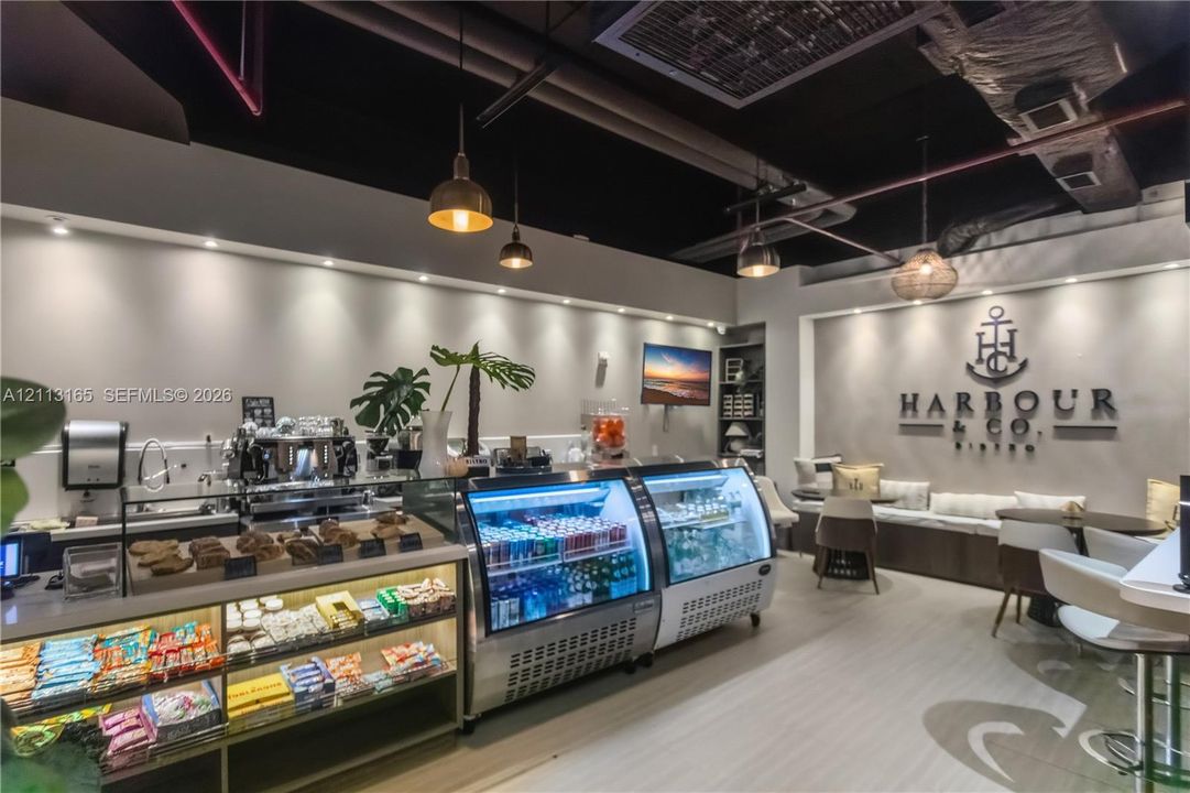 Commercial space featuring industrial-style exposed ceiling, recessed lighting, glass-front refrigerated display cases, light-toned wood-finish flooring, and built-in perimeter bench seating
