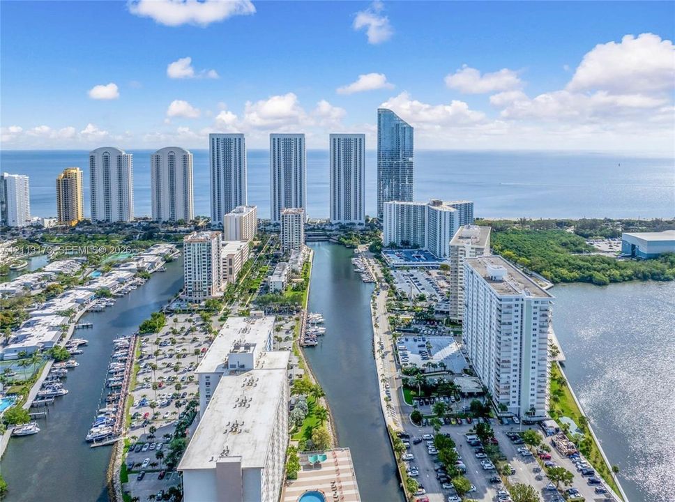 Aerial perspective of high-rise structures situated along an inlet, featuring direct water frontage, private boat docking, paved parking areas, and coastal surroundings