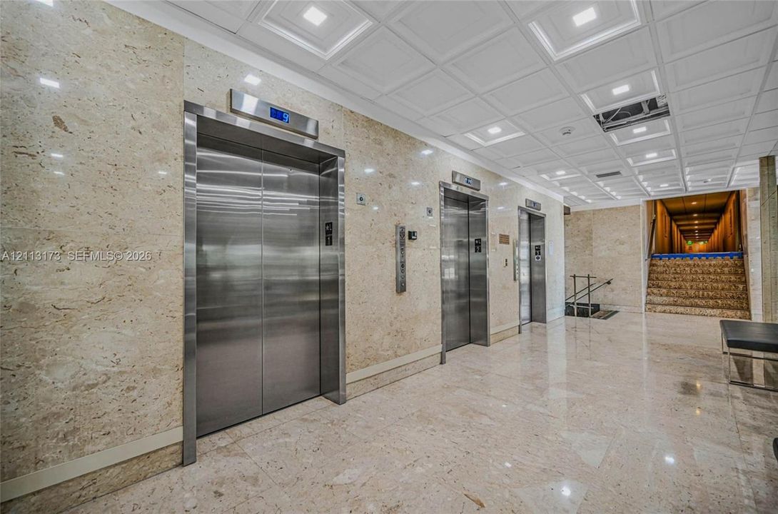 Polished stone wall cladding, stainless steel elevator doors, recessed ceiling lighting, coffered ceiling detail, and polished stone tile flooring