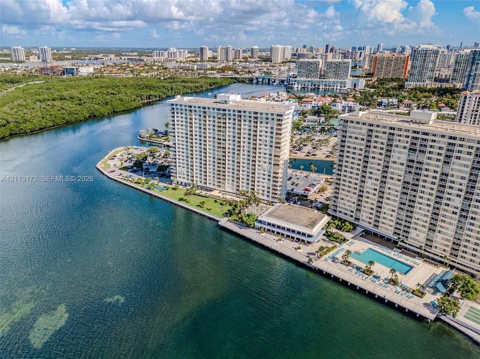 High-rise waterfront complex featuring a rectangular swimming pool, concrete seawall, paved parking areas, and manicured green spaces