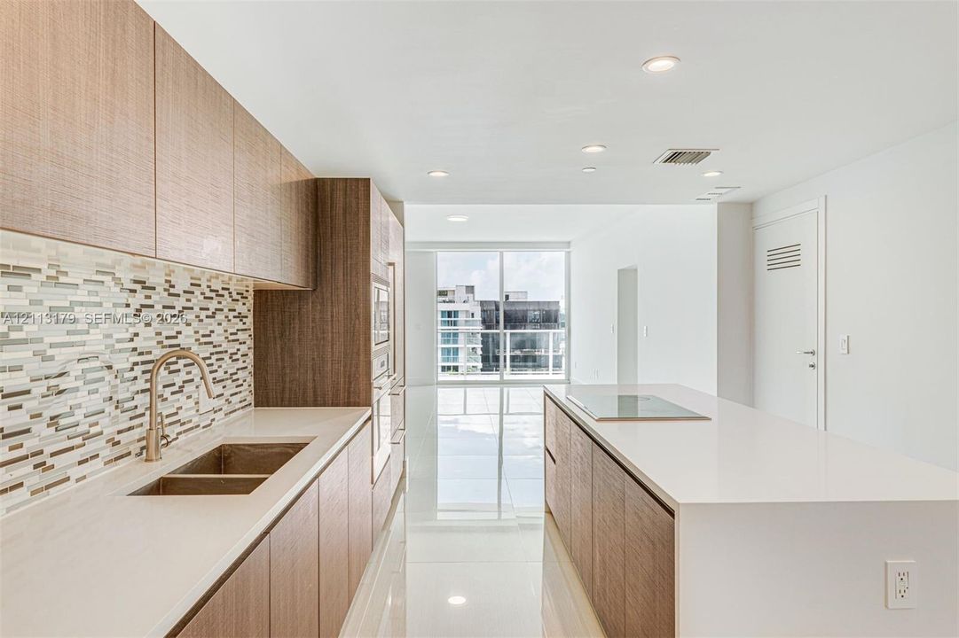 Kitchen featuring wood-finish cabinetry, stone-surface countertops, mosaic tile backsplash, integrated stainless steel appliances, and a central island with a flush-mount cooktop