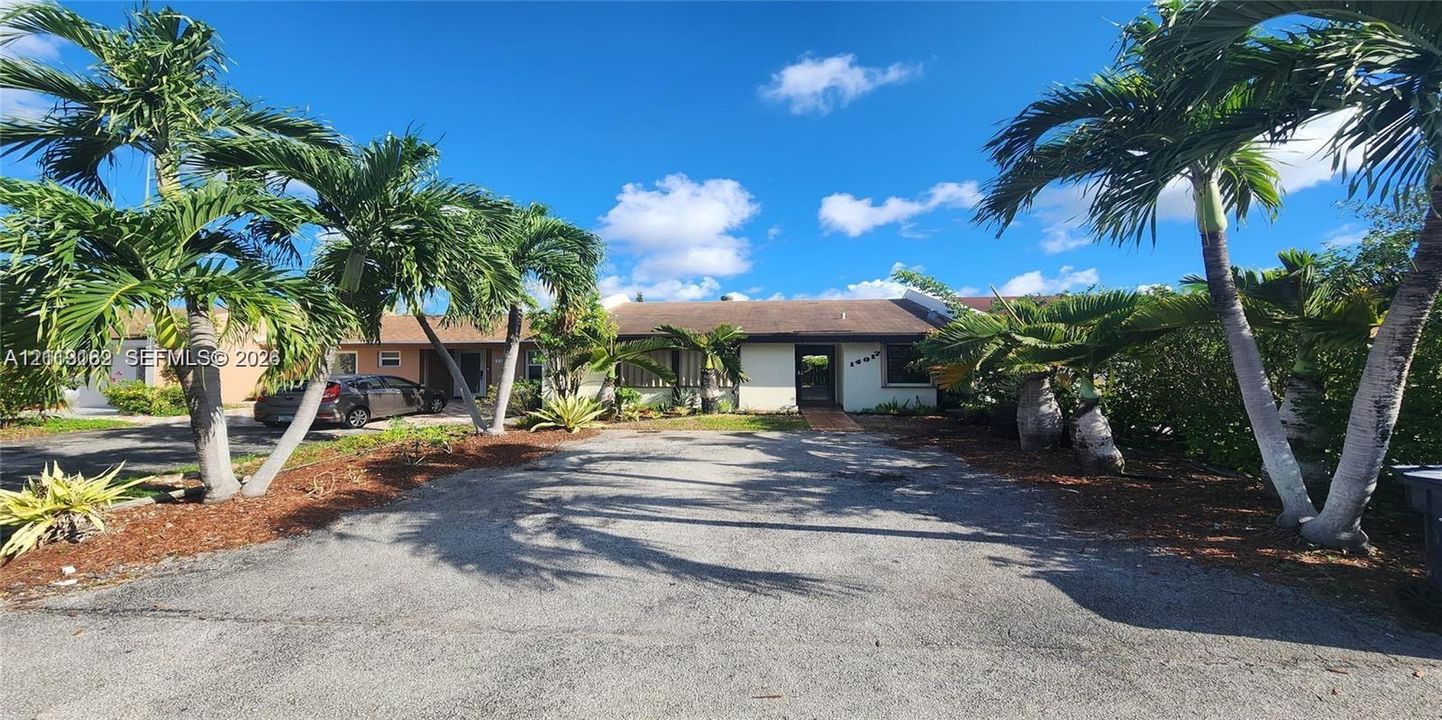 Single-story exterior featuring a light-toned stucco facade, pitched roofline, paved driveway, and established palm tree landscaping