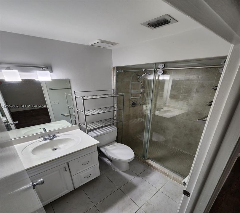 White vanity with an integrated sink, expansive mirror, walk-in shower with sliding glass doors, neutral tile flooring, and wall-mounted vanity lighting